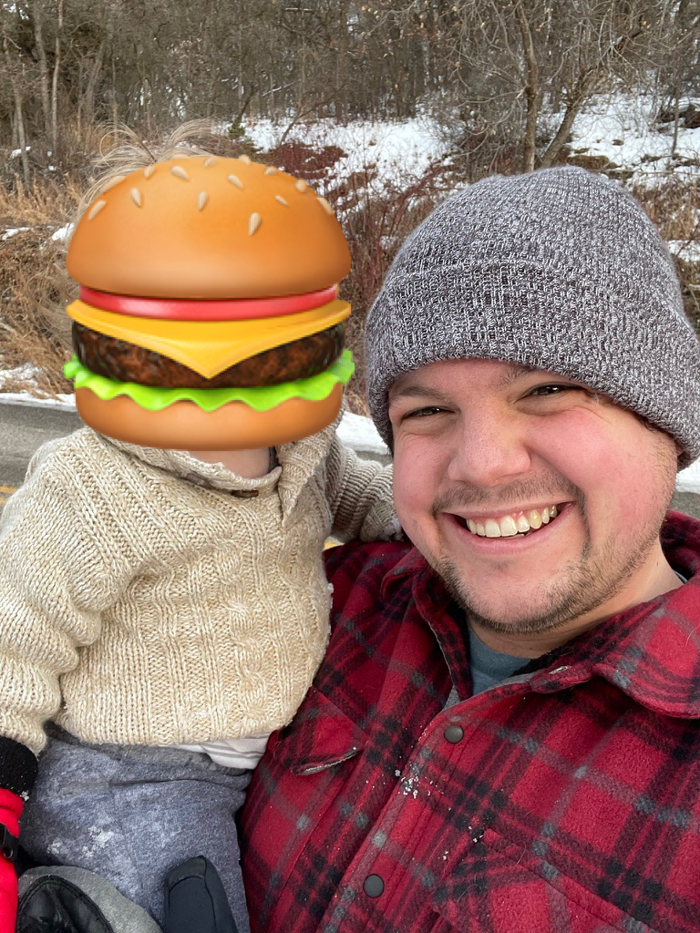 John Marc smiling outdoors with his young son, whose face is obscured by a burger emoji; bare trees and snow in the background.